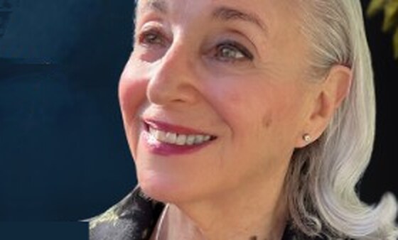 Naomi Ruth Lowinsky, smiling, with silver hair, green leaves, and a wooden sculpture behind her.